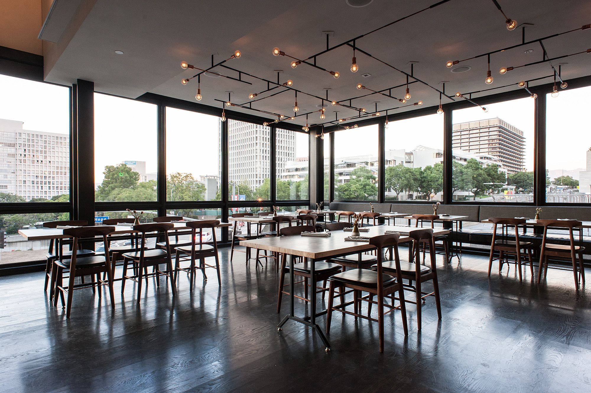 A modern, spacious dining area with large windows, wooden tables, and chairs. Overhead lighting features exposed bulbs. Cityscape visible outside.