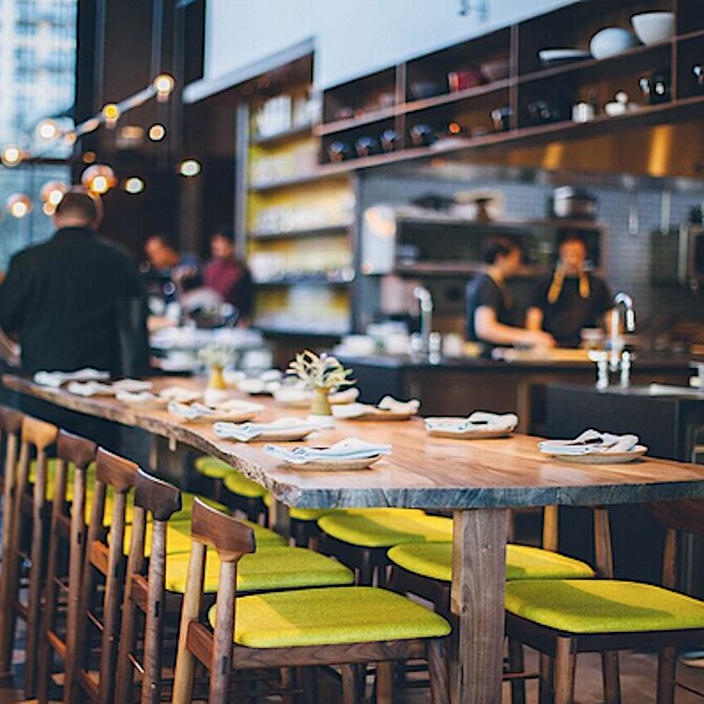 The WOOD High Stool (Bar Height) by Earl Home is a handcrafted wooden bar stool with a light finish, backrest, footrest, and simple design. It offers customizable upholstery to match any decor.