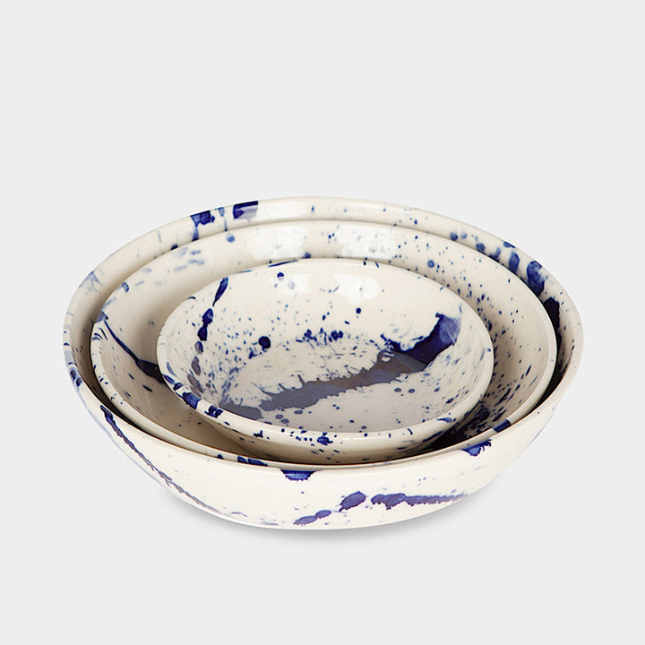 Earl Home's NESTING Bowls set includes three hand-thrown ceramic pieces with a blue splatter pattern on a plain background. Dishwasher and microwave safe, they blend artisanal design with daily functionality.