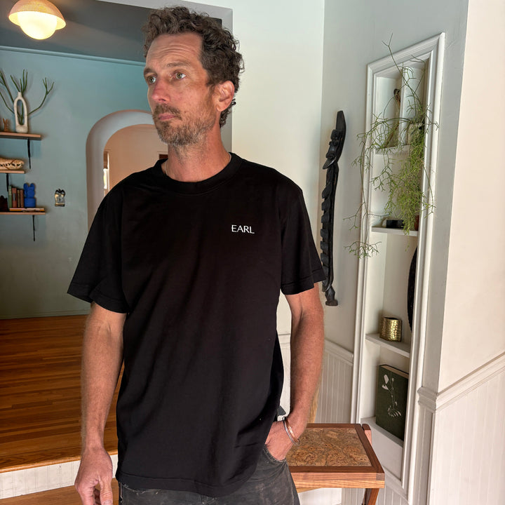 Man in a black t-shirt standing in a room with a staircase and light fixture.