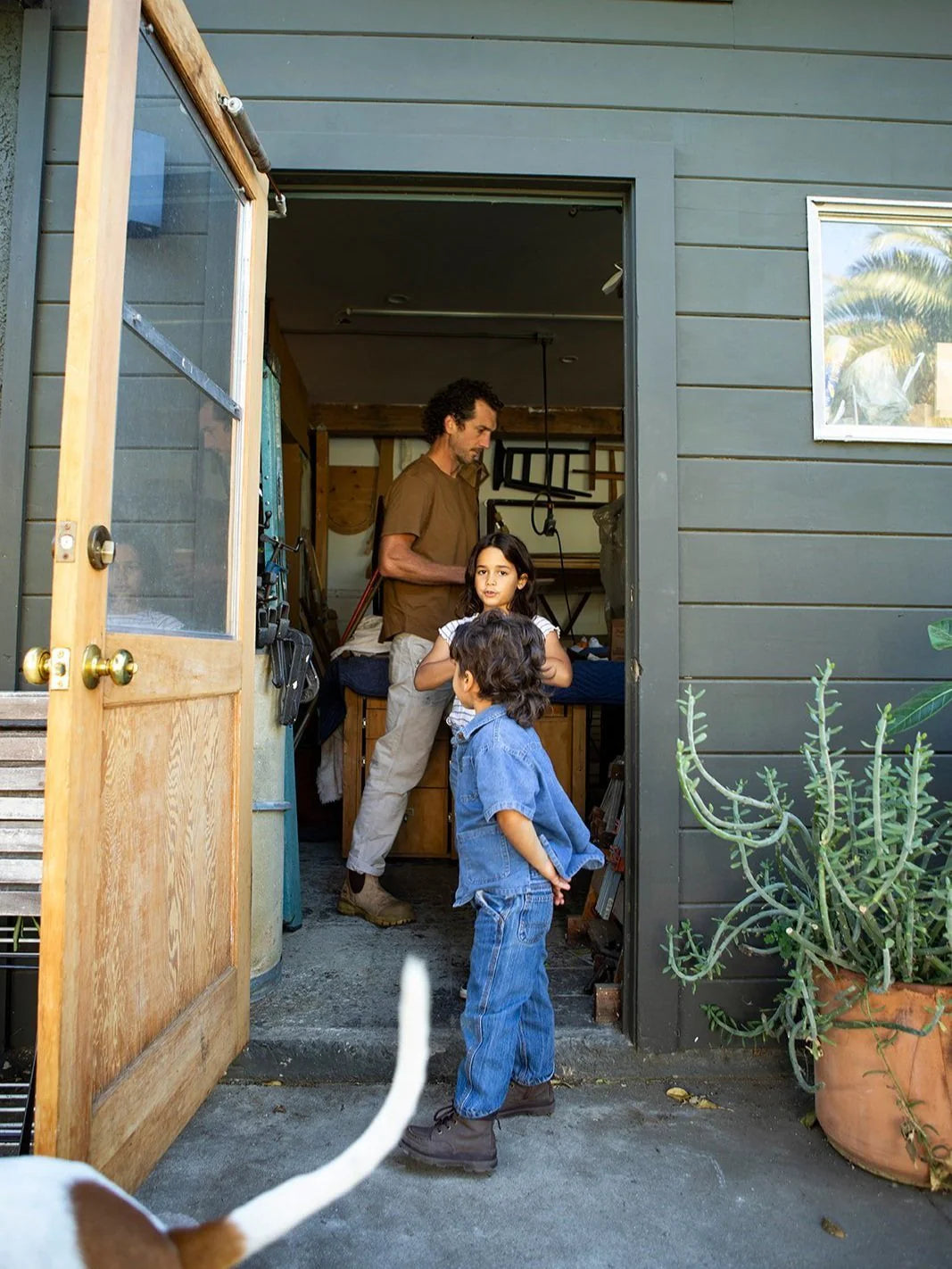 A man and two children stand near a partially open door of a house. A large plant is in a pot nearby, and a dog's tail is visible in the foreground.