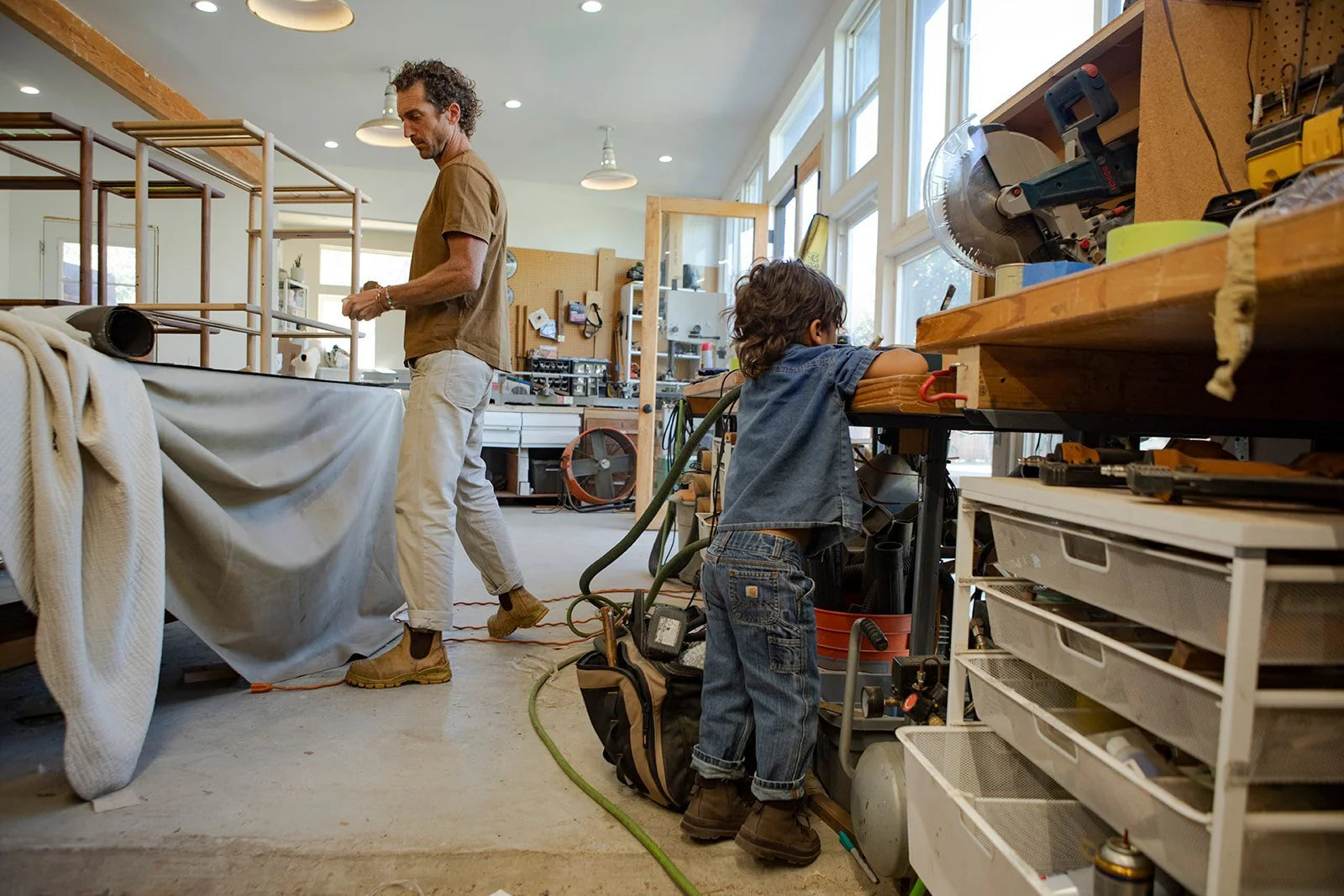 A man and a child are in a workshop. The man is standing near a workbench, while the child is leaning on another bench with tools around them.