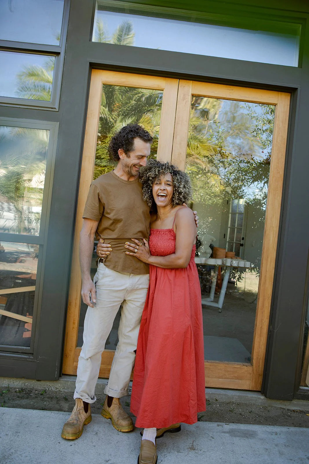 A man and woman stand smiling in front of a glass door. The woman is wearing a red dress, and the man is in a brown shirt and beige pants.