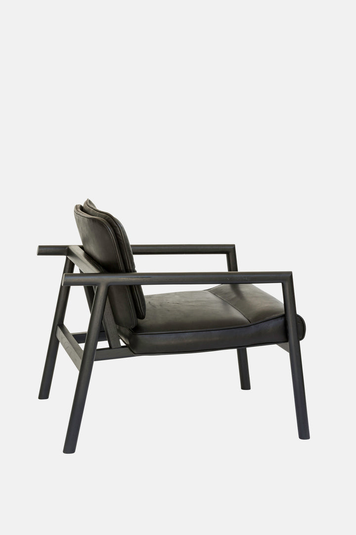 The MORESBY Lounge Chair by Earl Home is a modern, handmade piece featuring a sleek dark frame and a custom upholstered black leather cushion.