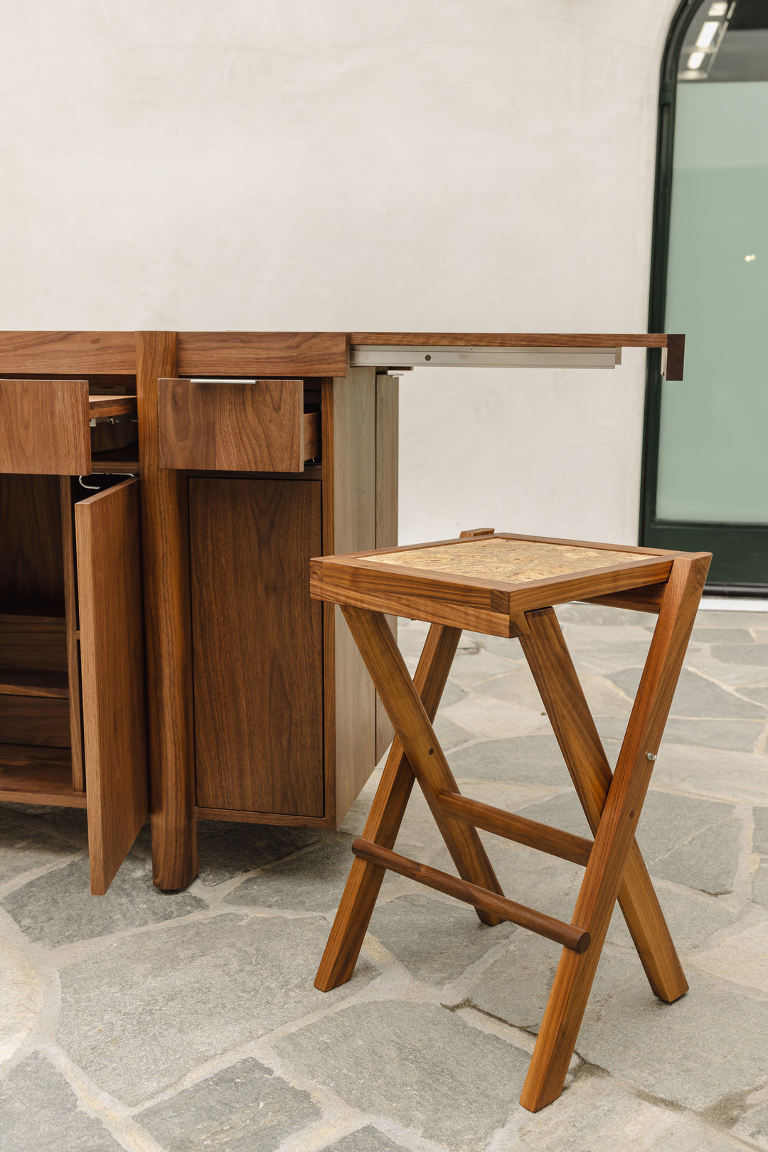 The LAPILO Folding Stool by Earl Home features an angular solid wood frame with a marbled cork seat and cross-legged design in a sleek finish.