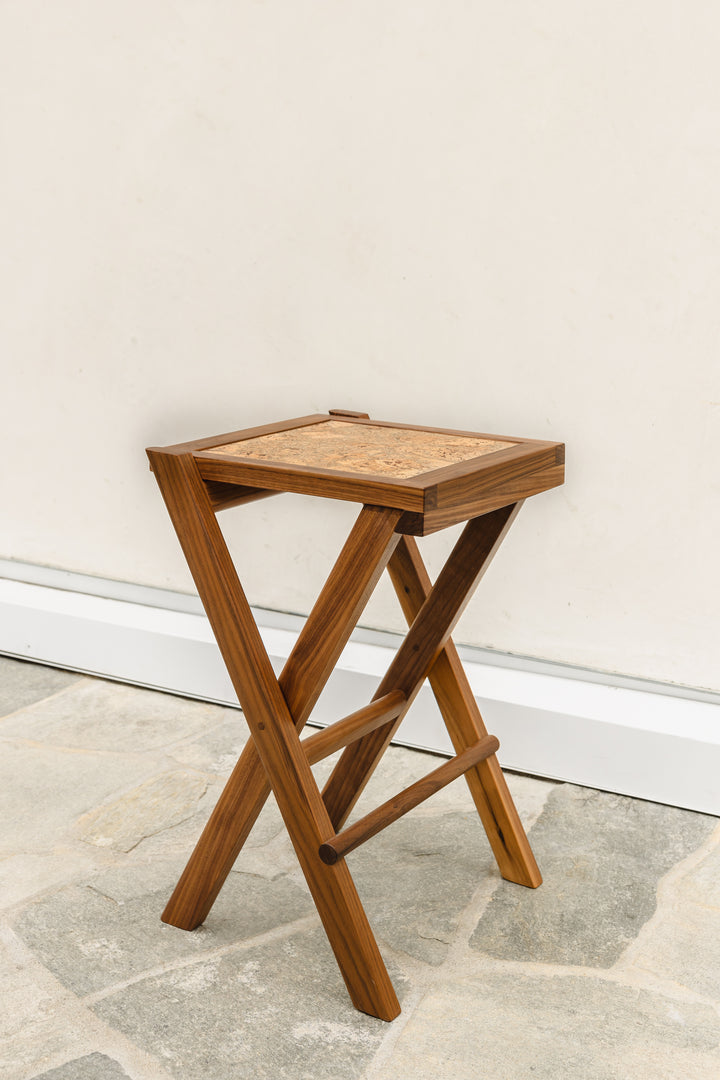 The LAPILO Folding Stool by Earl Home features an angular solid wood frame with a marbled cork seat and cross-legged design in a sleek finish.