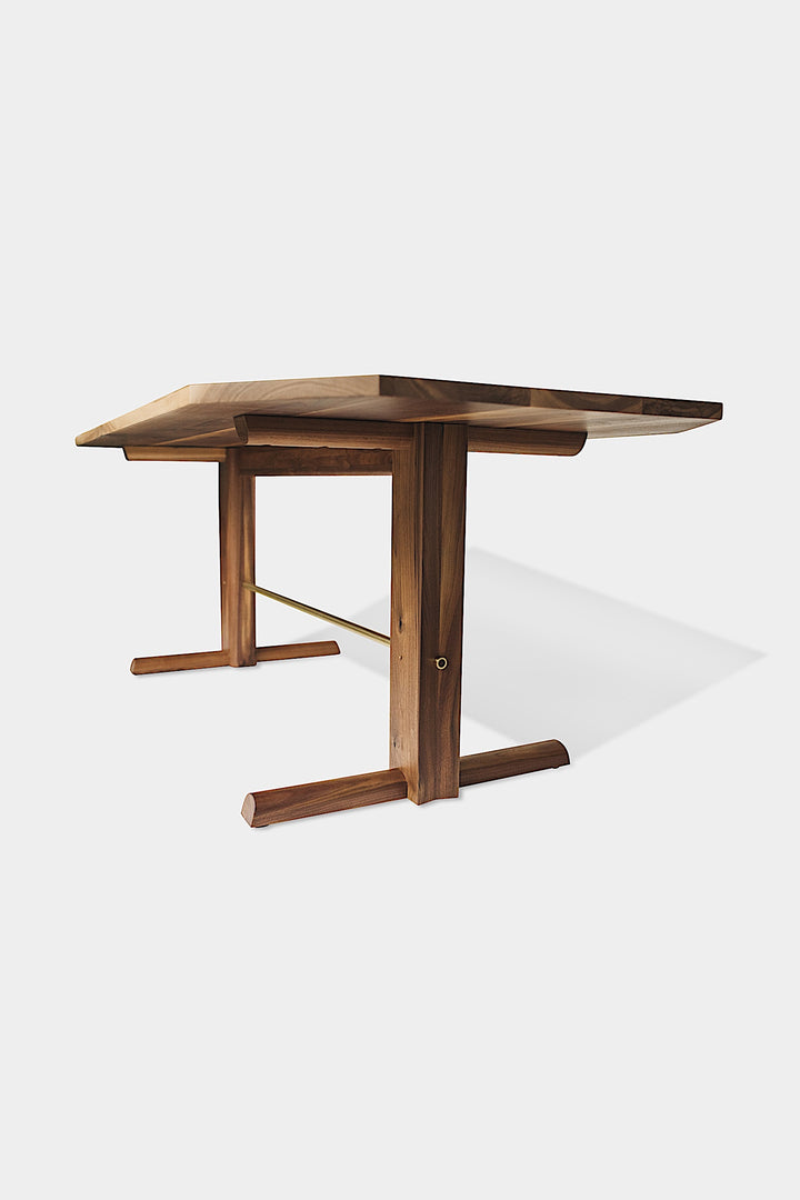 The BRIG Dining Table by Earl Home is a handmade piece featuring a rectangular solid wood top with angled corners, supported by two sturdy pedestal legs connected by a trestle base.