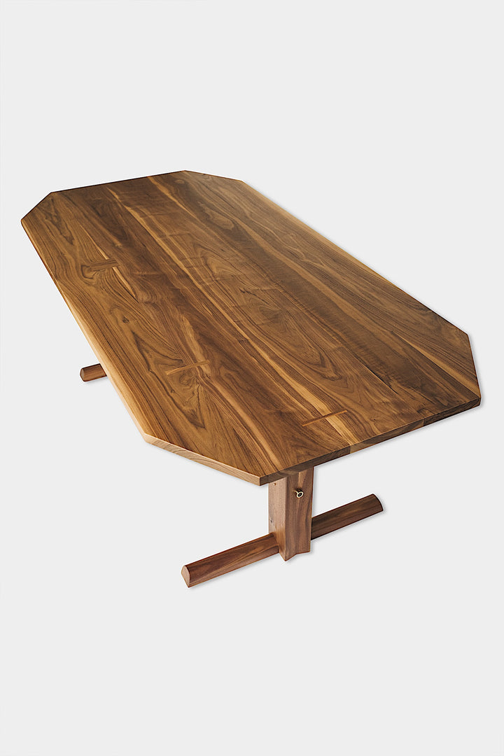 The BRIG Dining Table by Earl Home is a handmade piece featuring a rectangular solid wood top with angled corners, supported by two sturdy pedestal legs connected by a trestle base.