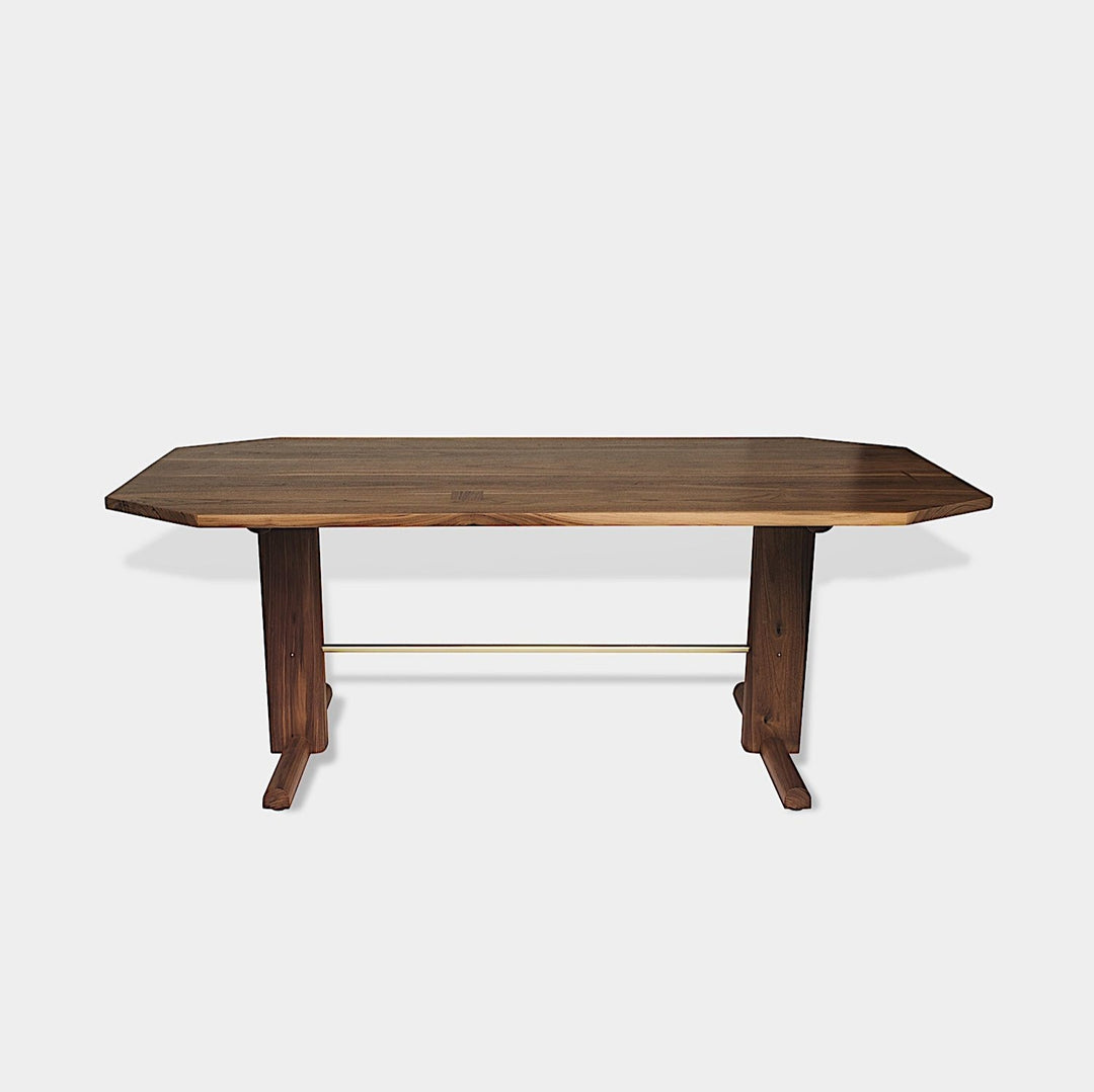 The BRIG Dining Table by Earl Home is a handmade piece featuring a rectangular solid wood top with angled corners, supported by two sturdy pedestal legs connected by a trestle base.