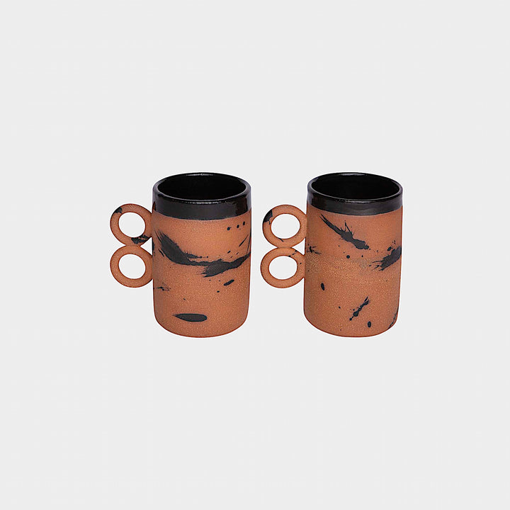 The Earl Home DOUBLE Handle Mug set includes two brown ceramic mugs, each featuring a unique round handle and white abstract patterns. Hand-thrown for artisanal charm, these mugs are perfect for those who appreciate the delicate artistry of ceramic craftsmanship.