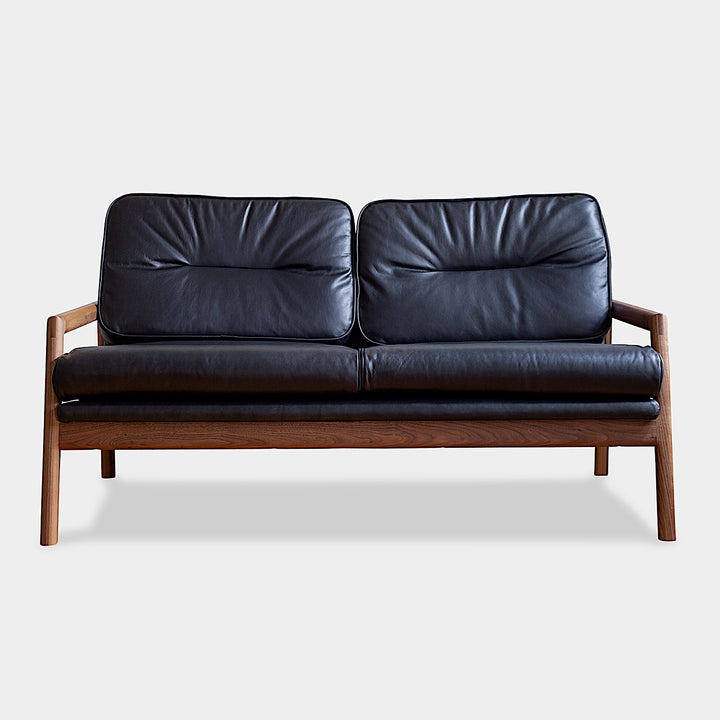 A front view of the MORESBY Love Seat by Earl Home, showcasing a walnut solid wood constructed frame and black leather upholstered cushions.