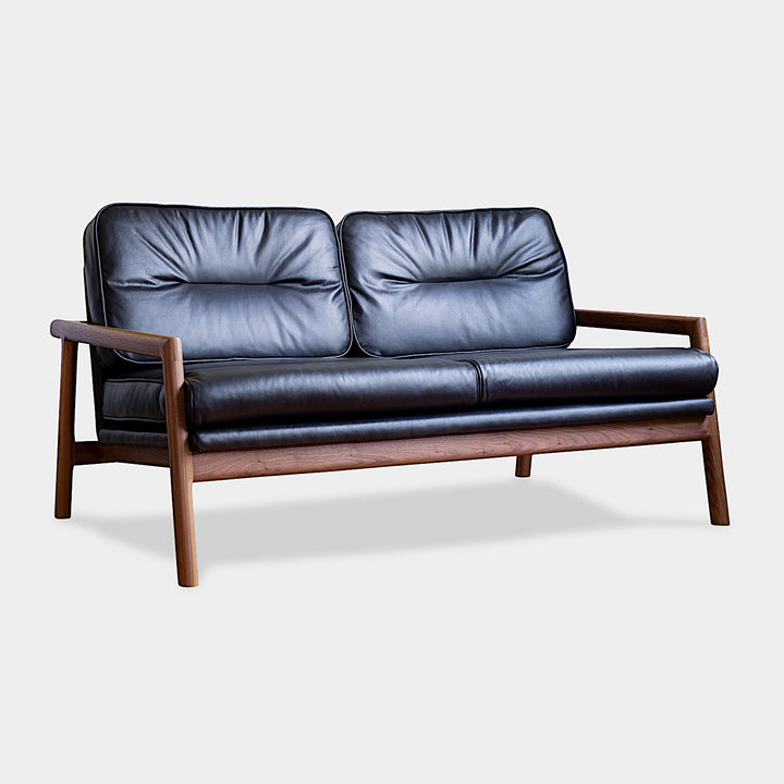 A front view of the MORESBY Love Seat by Earl Home, showcasing a walnut solid wood constructed frame and black leather upholstered cushions.