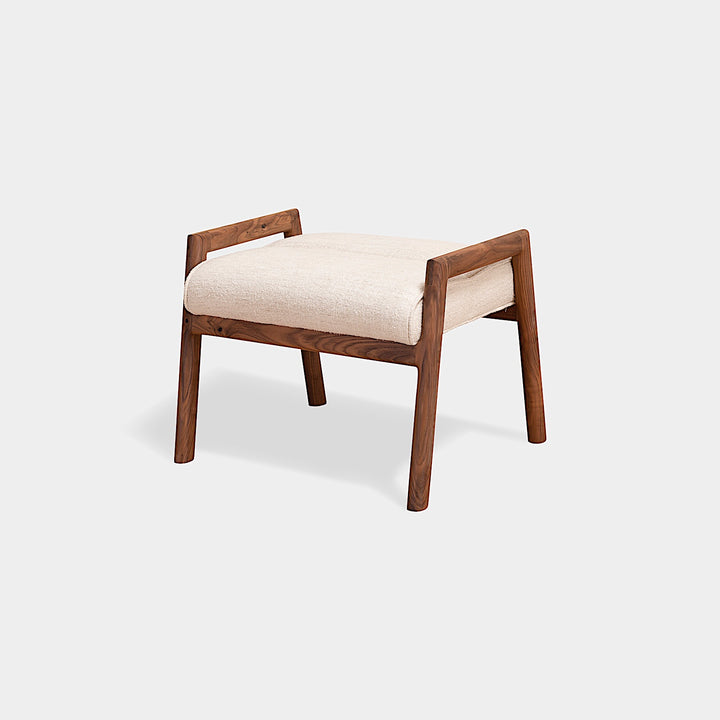 The MORESBY Ottoman by Earl Home is a handmade, custom order wooden stool with a light beige upholstered seat on four legs, set against a plain white background.