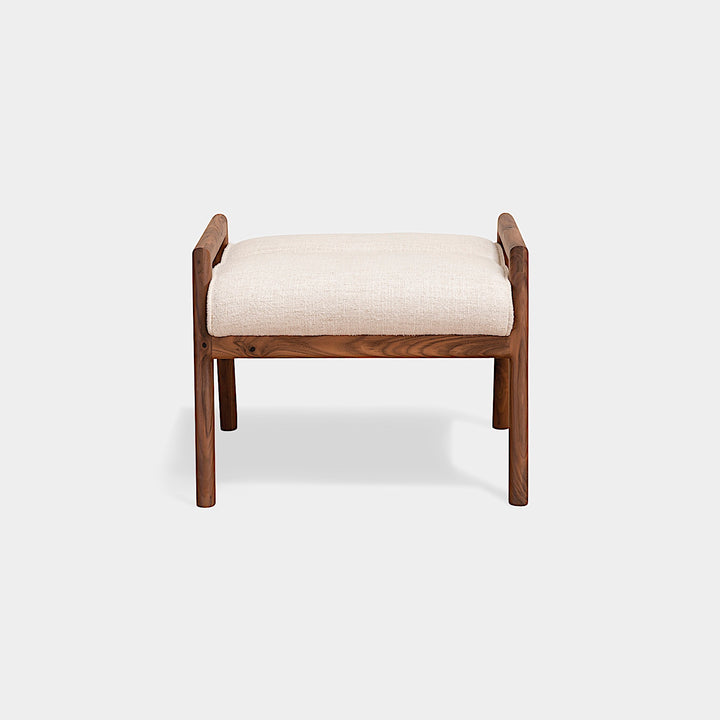 The MORESBY Ottoman by Earl Home is a handmade, custom order wooden stool with a light beige upholstered seat on four legs, set against a plain white background.