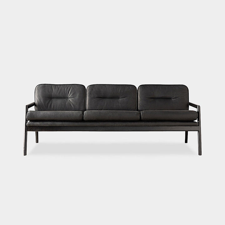 The MORESBY Sofa by Earl Home is a bespoke black leather piece, featuring custom upholstery, a metal frame, three cushions, and armrests. It's handmade to order for those who appreciate craftsmanship on a neutral background.