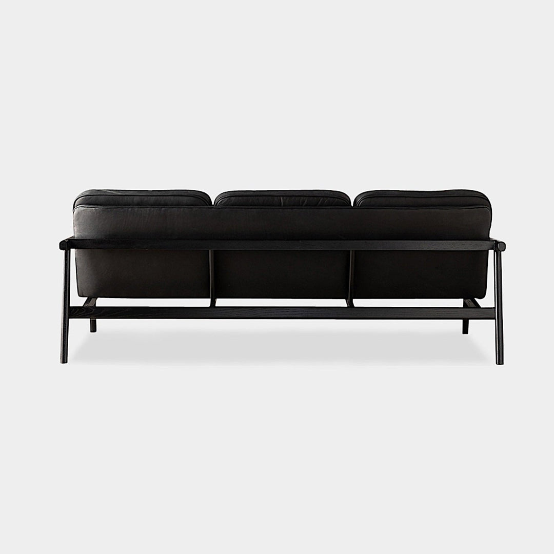 The MORESBY Sofa by Earl Home is a bespoke black leather piece, featuring custom upholstery, a metal frame, three cushions, and armrests. It's handmade to order for those who appreciate craftsmanship on a neutral background.