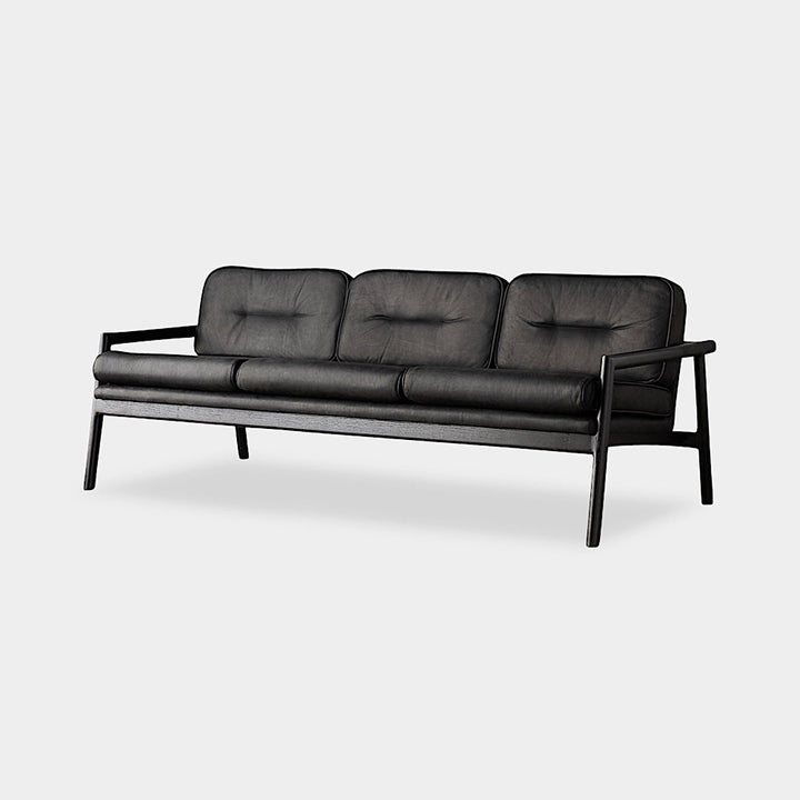 The MORESBY Sofa by Earl Home is a bespoke black leather piece, featuring custom upholstery, a metal frame, three cushions, and armrests. It's handmade to order for those who appreciate craftsmanship on a neutral background.