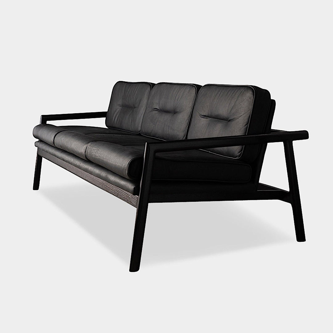 The MORESBY Sofa by Earl Home is a bespoke black leather piece, featuring custom upholstery, a metal frame, three cushions, and armrests. It's handmade to order for those who appreciate craftsmanship on a neutral background.