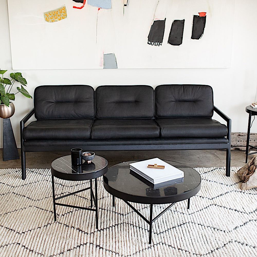 The MORESBY Sofa by Earl Home is a bespoke black leather piece, featuring custom upholstery, a black ebonized oak frame, three cushions, and armrests. It's handmade to order for those who appreciate craftsmanship in Sacha Strebe's old living room