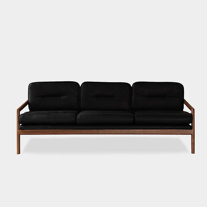 The MORESBY Sofa by Earl Home is a bespoke black leather piece, featuring custom upholstery, a metal frame, three cushions, and armrests. It's handmade to order for those who appreciate craftsmanship on a neutral background.