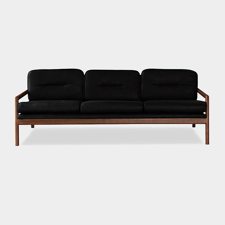 The MORESBY Sofa by Earl Home is a bespoke black leather piece, featuring custom upholstery, a metal frame, three cushions, and armrests. It's handmade to order for those who appreciate craftsmanship on a neutral background.