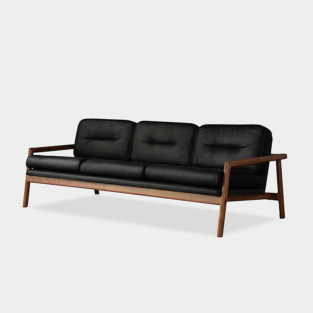 The MORESBY Sofa by Earl Home is a bespoke black leather piece, featuring custom upholstery, a metal frame, three cushions, and armrests. It's handmade to order for those who appreciate craftsmanship on a neutral background.