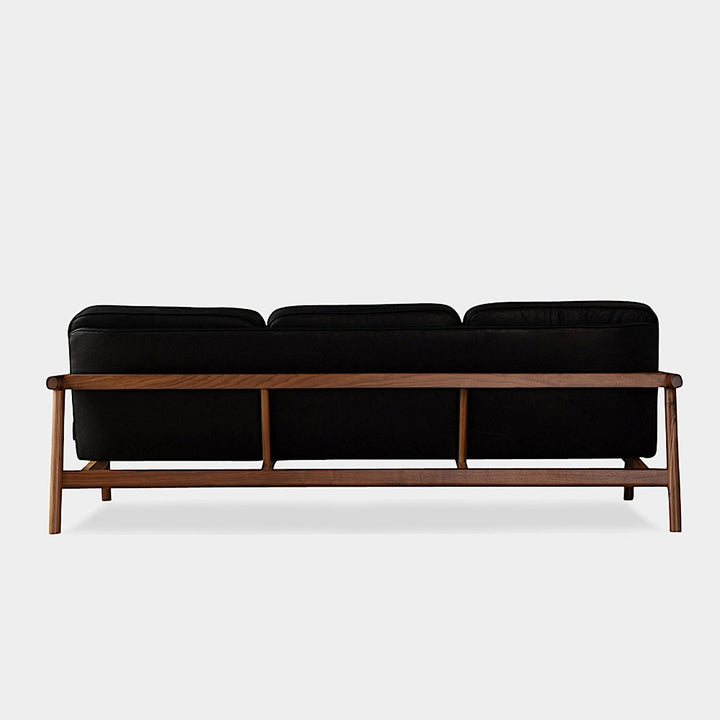 The MORESBY Sofa by Earl Home is a bespoke black leather piece, featuring custom upholstery, a metal frame, three cushions, and armrests. It's handmade to order for those who appreciate craftsmanship on a neutral background.