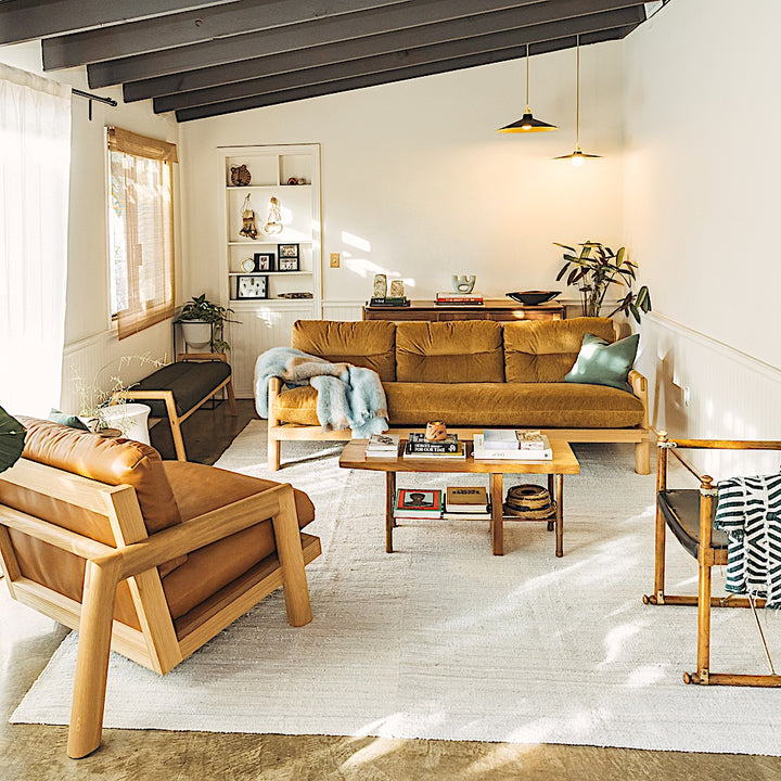 The White Oak TAMBU Sofa by Earl Home is a modern classic with a minimalist wooden frame of elegant White Oak, enhanced by three mustard cushions set against a white backdrop.