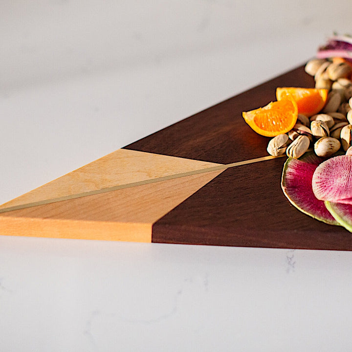 The TRIANGLE Cheese Board by Earl Home is a stylish centerpiece with a triangular design, lighter wood apex, darker base, brass and wood accents, and a central vertical division.