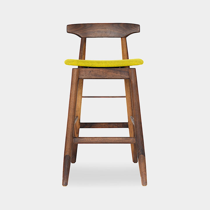 The WOOD High Stool (Bar Height) by Earl Home is a handcrafted wooden bar stool with a light finish, backrest, footrest, and simple design. It offers customizable upholstery to match any decor.