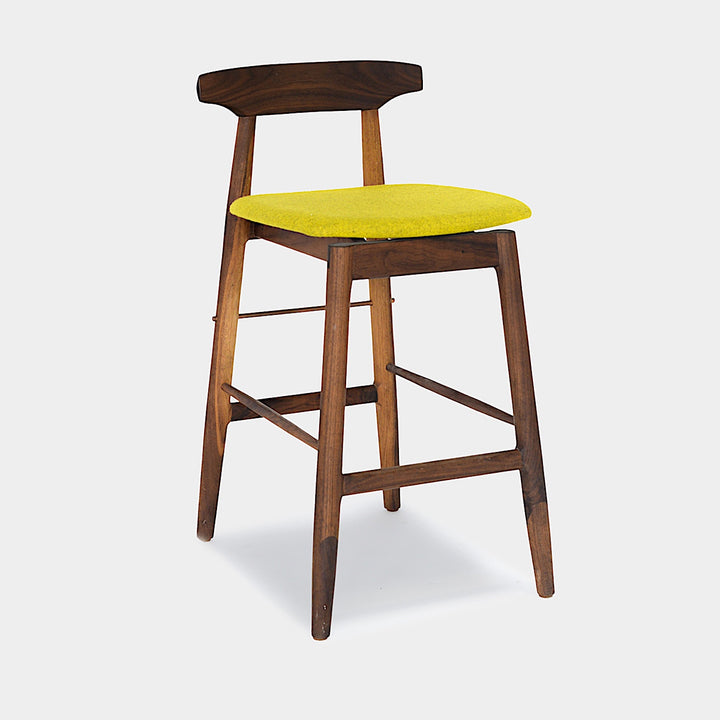 The WOOD High Stool (Bar Height) by Earl Home is a handcrafted wooden bar stool with a light finish, backrest, footrest, and simple design. It offers customizable upholstery to match any decor.