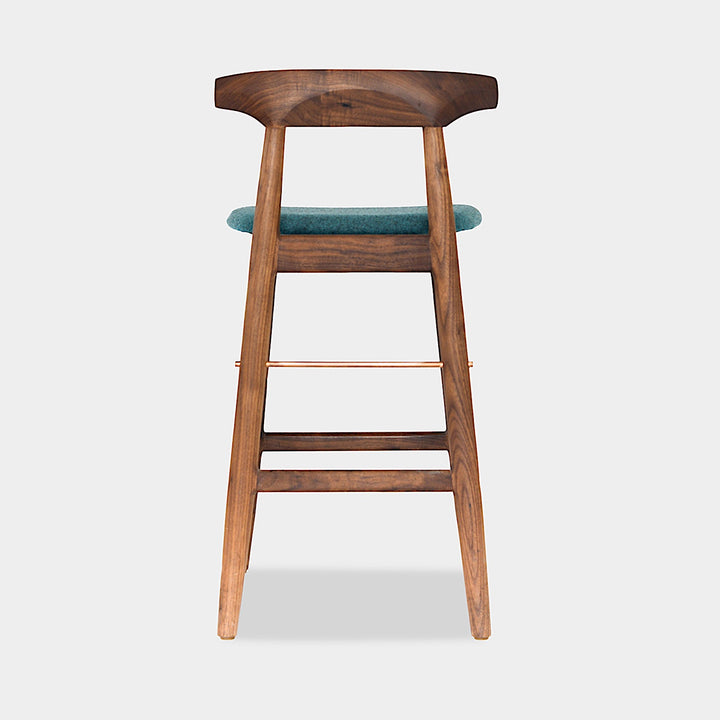 The WOOD High Stool (Bar Height) by Earl Home is a handcrafted wooden bar stool with a light finish, backrest, footrest, and simple design. It offers customizable upholstery to match any decor.