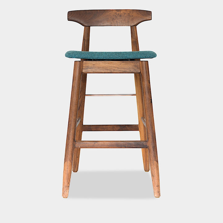 The WOOD High Stool (Bar Height) by Earl Home is a handcrafted wooden bar stool with a light finish, backrest, footrest, and simple design. It offers customizable upholstery to match any decor.