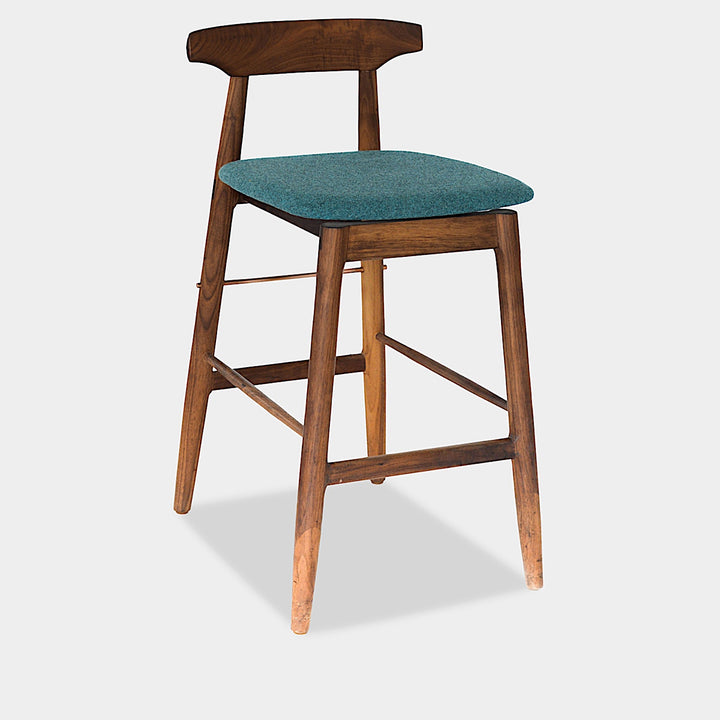 The WOOD High Stool (Bar Height) by Earl Home is a handcrafted wooden bar stool with a light finish, backrest, footrest, and simple design. It offers customizable upholstery to match any decor.