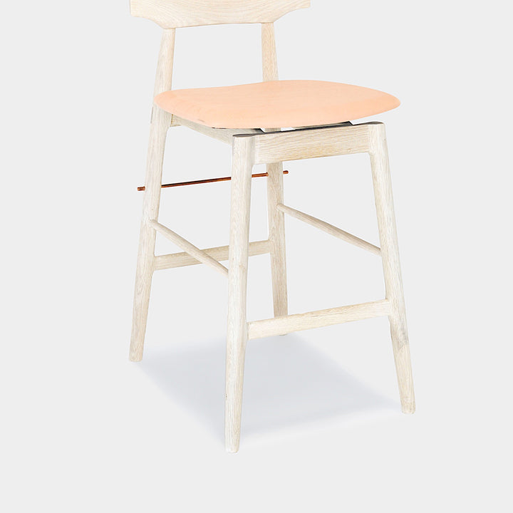 The WOOD High Stool (Bar Height) by Earl Home is a handcrafted wooden bar stool with a light finish, backrest, footrest, and simple design. It offers customizable upholstery to match any decor.