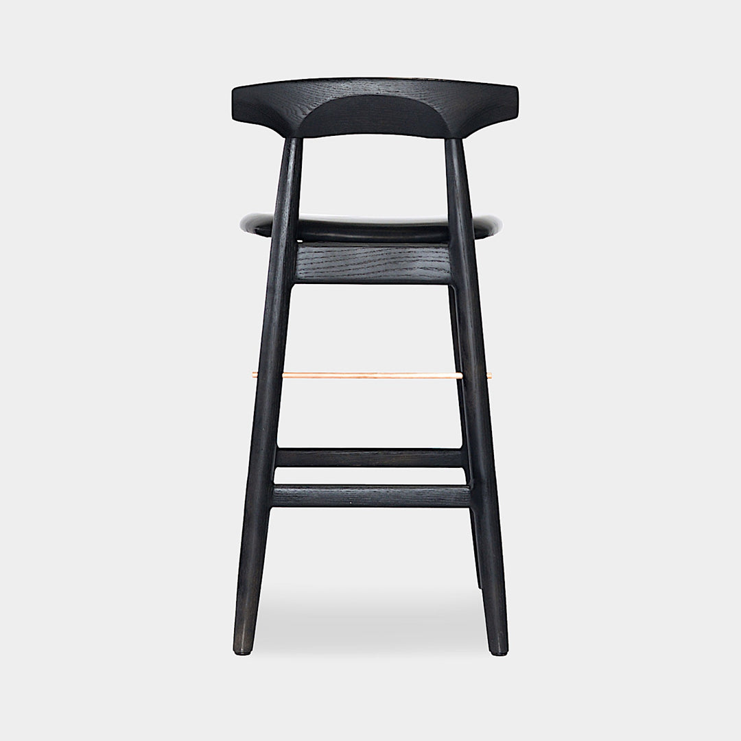 The WOOD High Stool (Bar Height) by Earl Home is a handcrafted wooden bar stool with a light finish, backrest, footrest, and simple design. It offers customizable upholstery to match any decor.