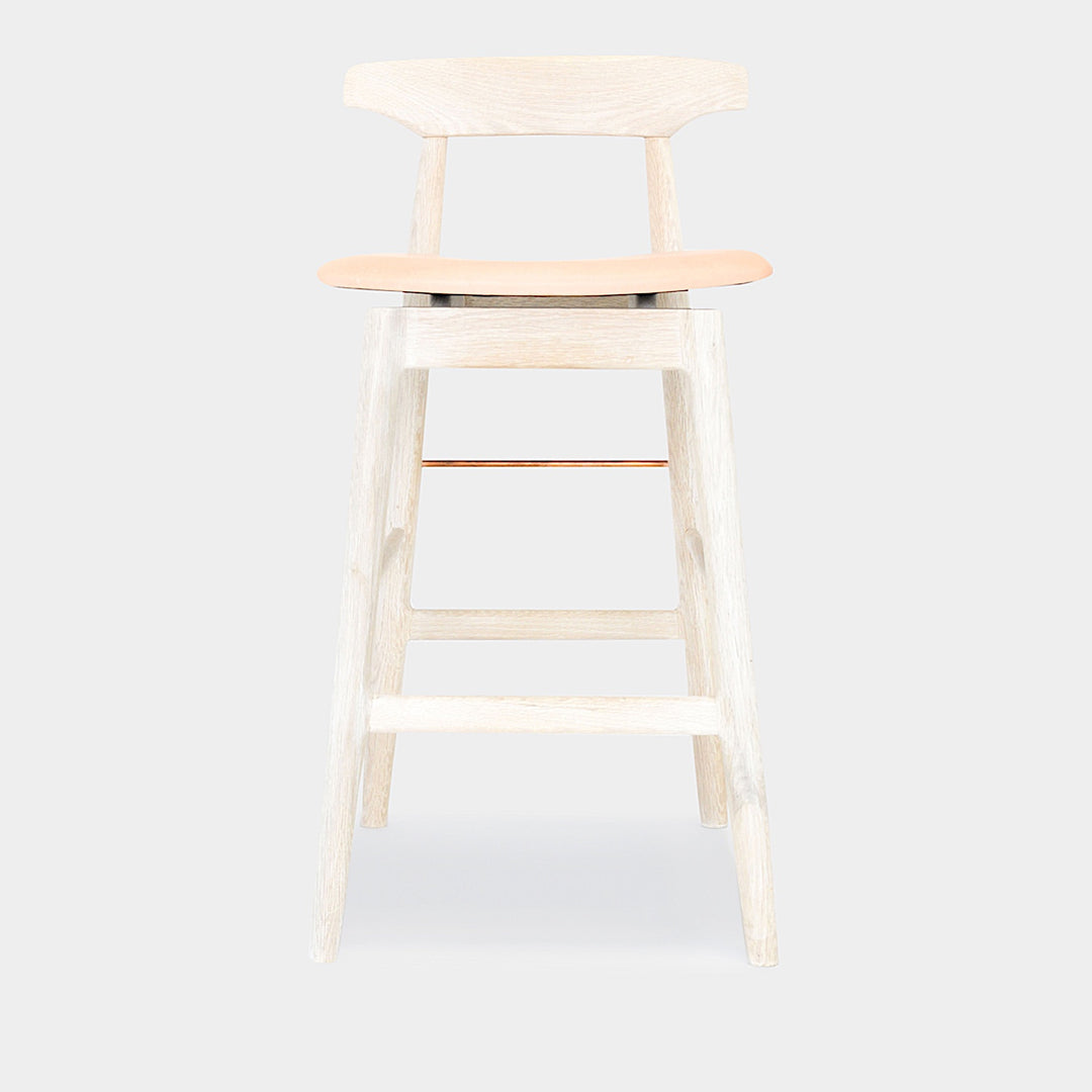 The WOOD High Stool (Bar Height) by Earl Home is a handcrafted wooden bar stool with a light finish, backrest, footrest, and simple design. It offers customizable upholstery to match any decor.