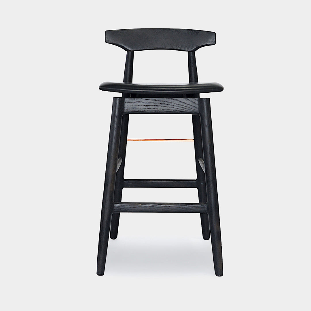 The WOOD High Stool (Bar Height) by Earl Home is a handcrafted wooden bar stool with a light finish, backrest, footrest, and simple design. It offers customizable upholstery to match any decor.