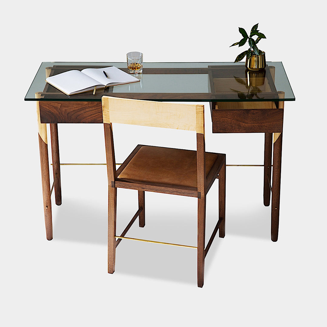 The WRITING Desk by Earl Home is an elegant glass desk with walnut and maple spliced wooden legs and a tempered glass top, Accompanied by a matching wooden chair. You can customize it to create your perfect workspace.