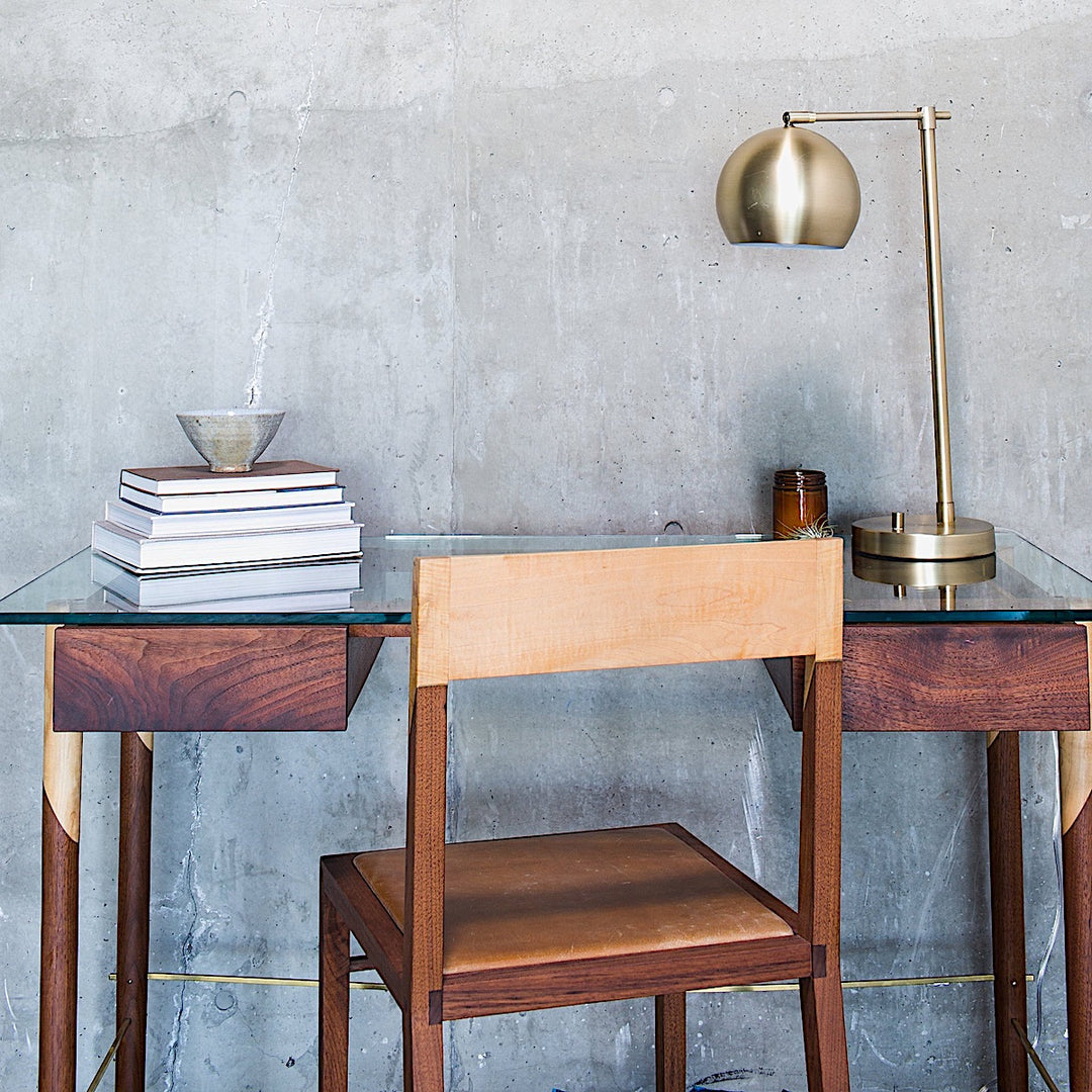 The WRITING Desk by Earl Home is an elegant glass desk with wooden legs and a natural edge, accompanied by a matching wooden chair. It includes an open notebook, a glass of water, and a potted plant for styling. You can customize it to create your perfect workspace.