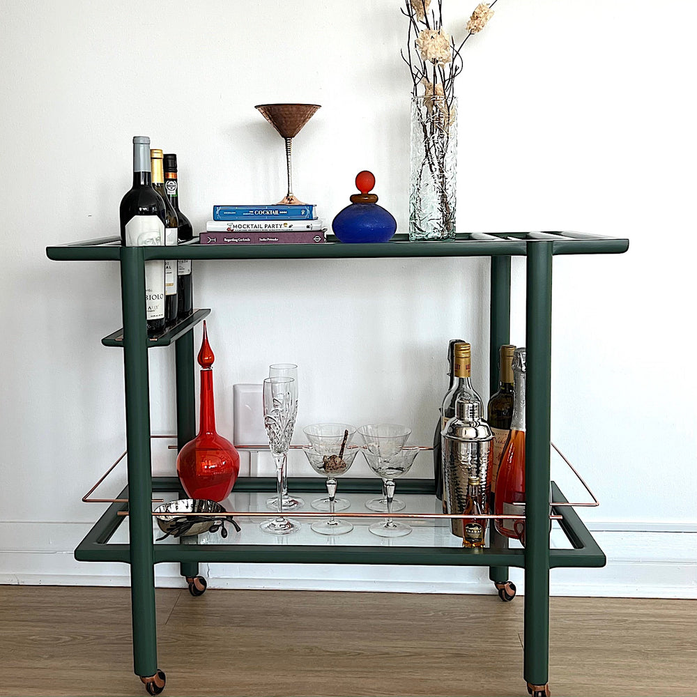 The Earl Home bar cart in deep green and copper showcases tempered glass panels and striking copper accents, complete with four wheels for easy movement.