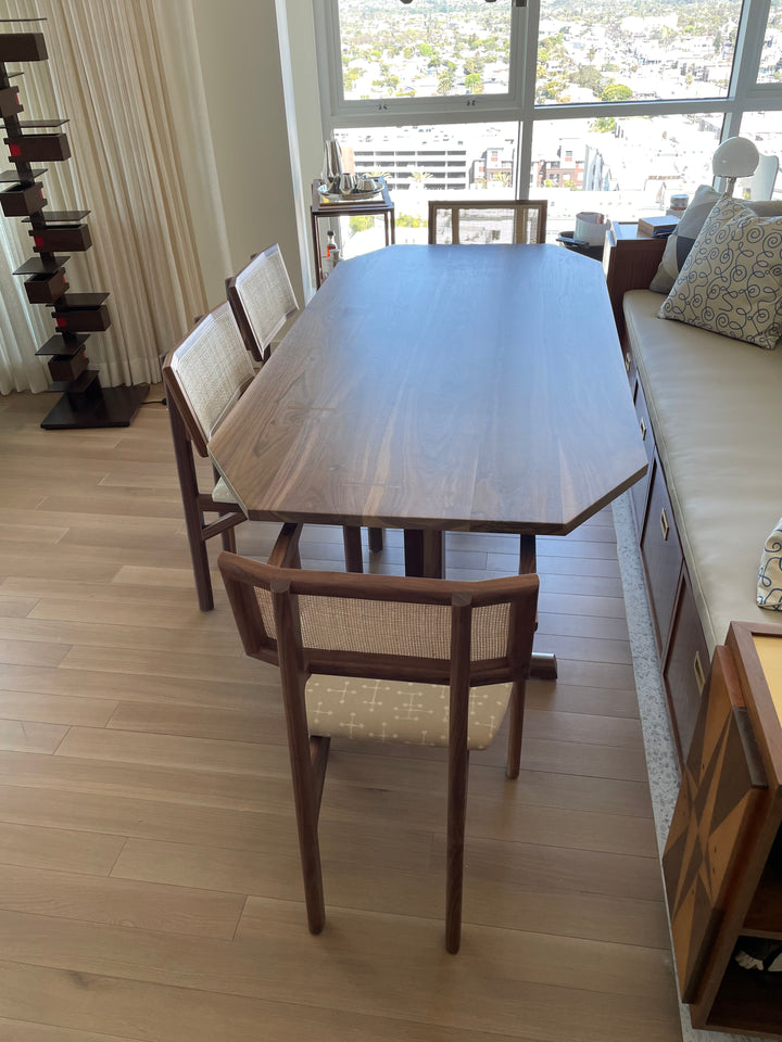 The BRIG Dining Table by Earl Home is a handmade piece featuring a rectangular solid wood top with angled corners, supported by two sturdy pedestal legs connected by a trestle base.