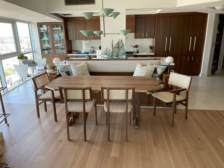 The BRIG Dining Table by Earl Home is a handmade piece featuring a rectangular solid wood top with angled corners, supported by two sturdy pedestal legs connected by a trestle base.