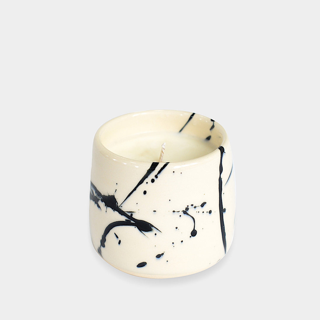 The Earl Home CANDLE is a white ceramic votive with a black splatter pattern, featuring an unlit cardamom-scented candle, set against a gray background.