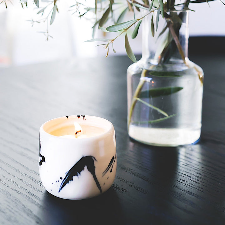 The Earl Home CANDLE is a white ceramic votive with a black splatter pattern, featuring an unlit cardamom-scented candle, set against a gray background.