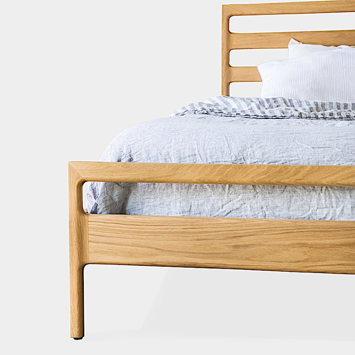 The LAMEL Bed by Earl Home is a hand-crafted bed frame with solid wood construction and a slatted headboard. It pairs beautifully with white pillows and a striped duvet, offering style and comfort. Available in custom bed sizes to fit your space perfectly.