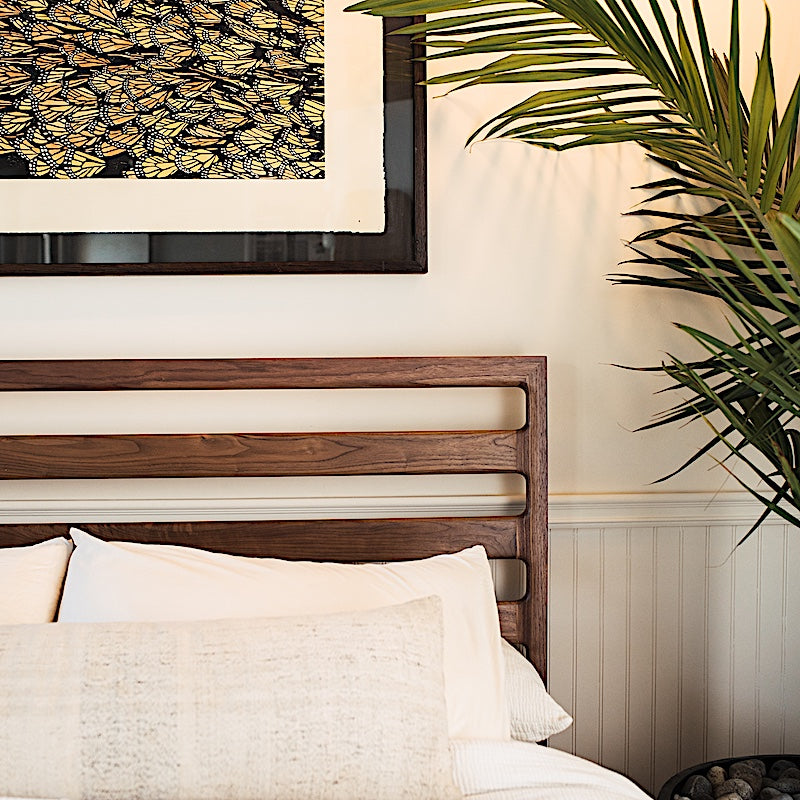 The LAMEL Bed by Earl Home is a hand-crafted bed frame with solid wood construction and a slatted headboard. It pairs beautifully with white pillows and a striped duvet, offering style and comfort. Available in custom bed sizes to fit your space perfectly.