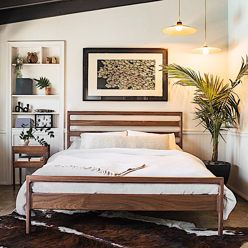 The LAMEL Bed by Earl Home is a hand-crafted bed frame with solid wood construction and a slatted headboard. It pairs beautifully with white pillows and a striped duvet, offering style and comfort. Available in custom bed sizes to fit your space perfectly.