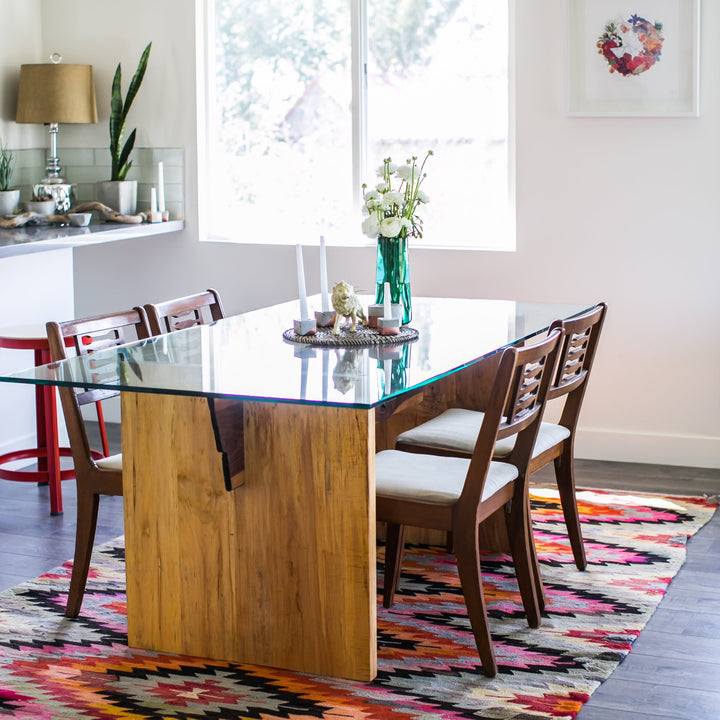 The LUXE Dining Table by Earl Home features a polished glass top and a wooden base with natural wood grain and geometric lines. Solid maple uprights with a walnut spine.