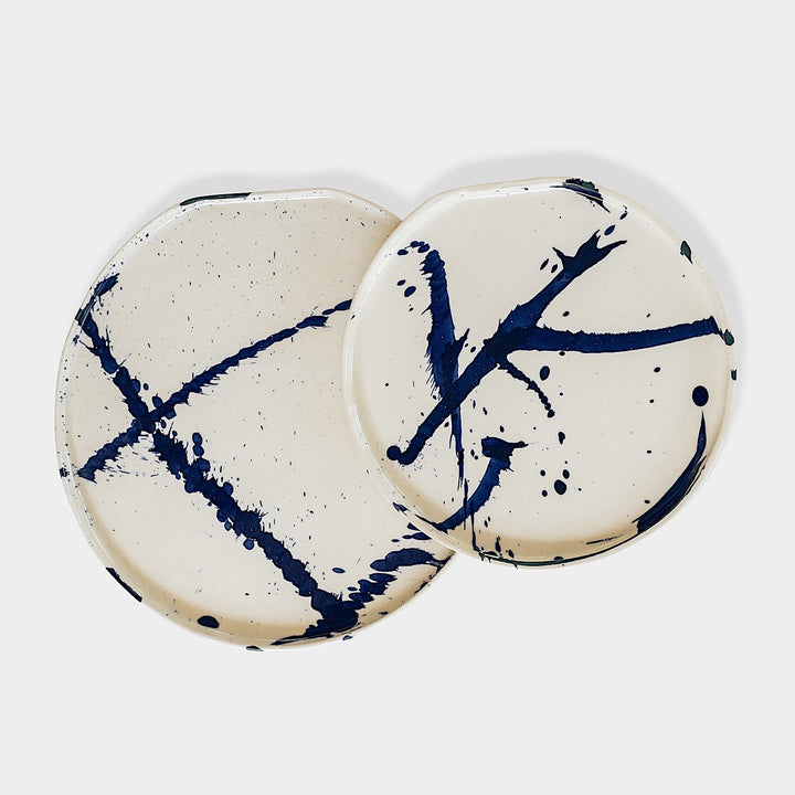 The Earl Home PLATES feature a round ceramic design with a white base and two intersecting dark blue splatter patterns. They are both dishwasher and microwave safe.